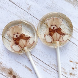 May include: Two round lollipops with a brown teddy bear design on a white background. The lollipops have gold glitter embedded in the clear candy.