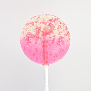 May include: A round lollipop featuring a gradient design that transitions from a clear, translucent top to a solid, vibrant pink bottom. The candy surface is embedded with small, sparkling pink and gold flecks throughout, attached to a white stick.