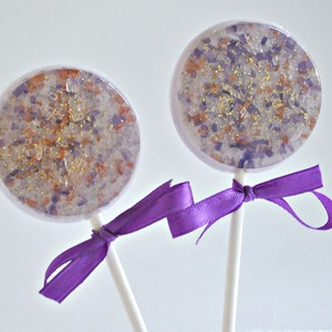 May include: Two round lollipops with clear candy, filled with purple, orange, and gold sprinkles. Each lollipop is attached to a white stick and tied with a purple ribbon. The lollipops are on a white background.