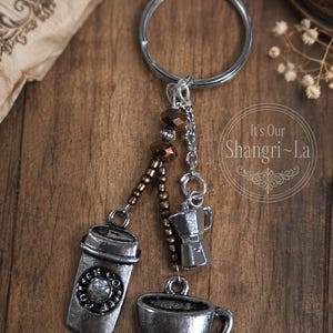May include: A silver-toned metal keychain featuring three coffee-themed charms: a travel cup with the text COFFEE, a moka pot, and a coffee cup on a saucer. The charms are attached to a circular ring with small brown and silver beads on chains.