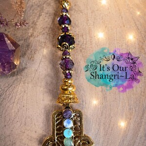 May include: A gold-toned metal keychain featuring a Hamsa hand charm adorned with seven small, vertically aligned circular stones in rainbow colors. Above the charm, a series of dark purple faceted beads are separated by gold-toned spacers.