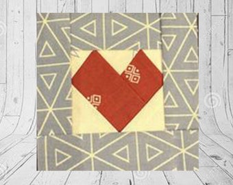 May include: A fabric block with a red heart design on a cream-colored square, set against a gray background with a geometric pattern. The heart has a small square pattern on it. The background has a triangle pattern.