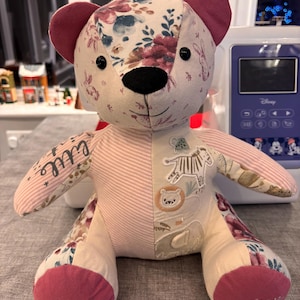 May include: A patchwork teddy bear with floral and animal print fabric. The bear features a pink and white striped torso, dark pink ears and feet, and the name "Annabelle" embroidered on one foot. The bear is on a grey surface.