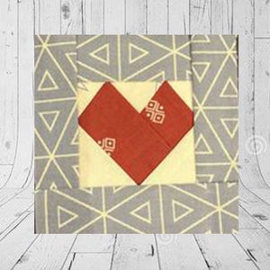 May include: A fabric block with a red heart design on a cream-colored square, set against a gray background with a geometric pattern. The heart has a small square pattern on it. The background has a triangle pattern.