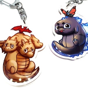 May include: Two keychains featuring cartoon characters. The first keychain features a three-headed dragon with red wings. The second keychain features a gray Godzilla-like monster with a red butterfly on its head.
