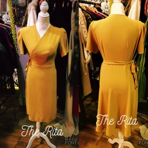 May include: A mustard yellow wrap dress with short sleeves and a tie at the waist. The dress is on a white mannequin and the text "The Rita" is written below the dress.