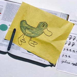 May include: A green crayon drawing of a duck on a yellow piece of paper. The paper is on top of an open textbook with a blue crayon next to it.