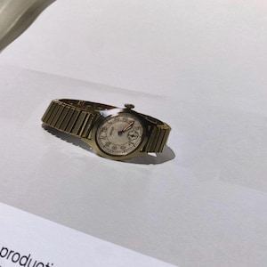 May include: A gold-toned wristwatch with a white face and black hands. The watch has a metal band.