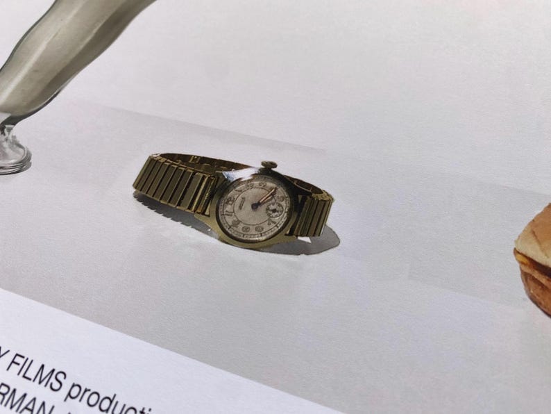 May include: A gold-toned wristwatch with a white face and black hands. The watch has a metal band.