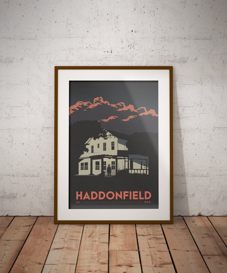 May include: A framed poster with a dark background featuring a white house with a silhouette of a person standing in front of it. The sky is a dark grey with pink clouds. The text "HADDONFIELD IL USA" is printed in red at the bottom of the poster.
