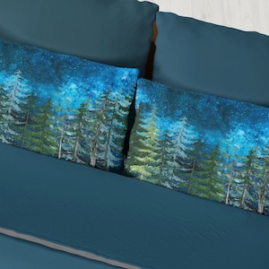 May include: Two blue pillowcases with a watercolor design of a forest at night with a starry sky. The pillowcases are on a blue bedspread.