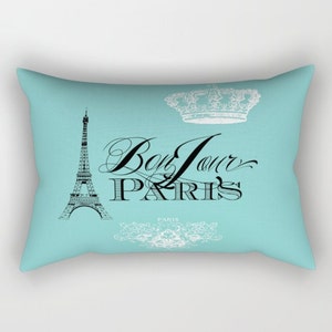 K&ouml;nnte beinhalten: Ein t&uuml;rkisfarbenes Kissen mit einem schwarz-wei&szlig;en grafischen Design, das den Eiffelturm, eine Krone und den Text "Bon Jour Paris" zeigt.