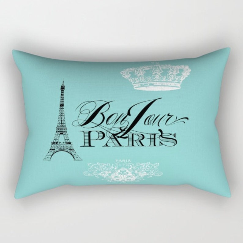 K&ouml;nnte beinhalten: Ein t&uuml;rkisfarbenes Kissen mit einem schwarz-wei&szlig;en grafischen Design, das den Eiffelturm, eine Krone und den Text "Bon Jour Paris" zeigt.