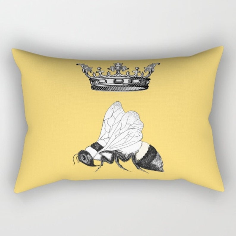 Pu&ograve; includere: Un cuscino rettangolare giallo con un'illustrazione in bianco e nero di un'ape e una corona. L'ape ha un addome a strisce e ali delicate. La corona &egrave; ornata, aggiungendo un tocco regale al design.