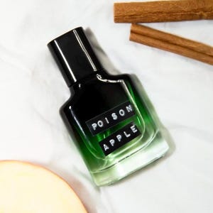 May include: A green glass bottle of perfume with a black cap and a white label that says "POISON APPLE". The bottle is sitting on a white surface with a cinnamon stick and a slice of apple.