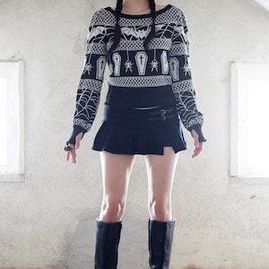 May include: A black and white patterned sweater with a coffin, spider, and bat design. The sweater is worn with a black mini skirt and black knee-high boots.