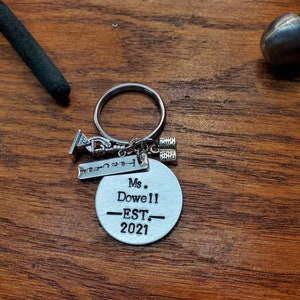 May include: Silver keychain with a round charm that says "Ms. Dowell -EST. 2021" and a small charm shaped like a graduation cap.