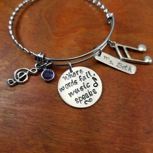 May include: Silver charm bracelet with a blue gem, a treble clef charm, and a charm with the text "Where words fail, music speaks". The bracelet also has a tag with the text "Ms. Beth" and a musical note charm.