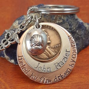 Pode incluir: Chaveiro de prata e cobre com um símbolo médico, um pequeno amuleto de chapéu de prata e um centavo americano de 1950. O chaveiro tem as palavras "John Frazier" e "fueled by fire driven by courage" gravadas.