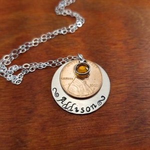May include: A silver chain necklace with a pendant featuring a 2005 US penny with a small amber gemstone on top and a silver disc with the name "Addison" engraved on it.