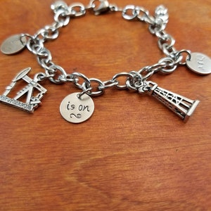 May include: Silver charm bracelet with various charms, including a miniature oil derrick, a tower, and round charms with engraved text. The bracelet is made of linked chain and has a lobster clasp.