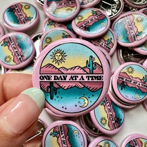 Pode incluir: Uma coleção de broches de botão rosa com um design de paisagem desértica. Os broches apresentam um cacto, montanhas, um sol, uma lua crescente e estrelas. O texto "ONE DAY AT A TIME" está impresso nos broches.
