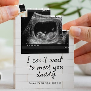 May include: A white building block construction featuring a black and white ultrasound image at the top. The text reads, "I can't wait to meet you daddy" and "Love from the bump x" in black lettering. The structure is composed of interlocking plastic bricks.