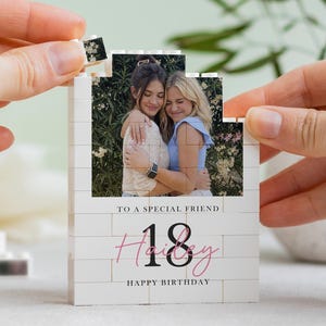May include: A white brick-style block with a photo of two women hugging. The block reads "TO A SPECIAL FRIEND" and "18 Happy Birthday" in pink and black text. A hand is placing a small block on top.