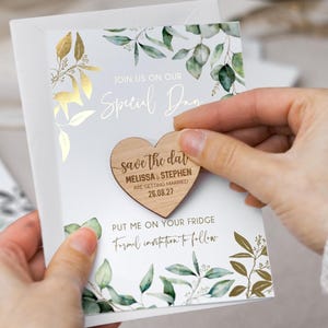 May include: A white save the date card featuring green eucalyptus leaves and gold foil accents. The card includes the text Join us on our special day and Put me on your fridge. A heart-shaped wooden magnet is attached, engraved with save the date, Melissa & Stephen are getting married, and 26.08.27.