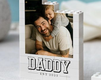 Puede incluir: Un marco de fotos rectangular construido con bloques de construcción de plástico blanco entrelazados. El marco muestra una fotografía de una persona con un niño y presenta el texto impreso en negro DADDY EST 2023 en la parte inferior.