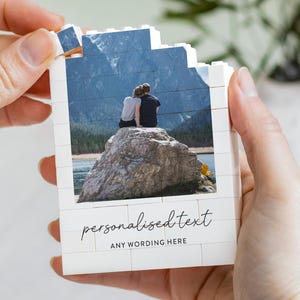 Può includere: Una costruzione rettangolare in mattoncini bianchi con una fotografia stampata di due persone sedute su una grande roccia davanti a una montagna e un lago. La parte inferiore mostra il testo "personalized text" e "ANY WORDING HERE".
