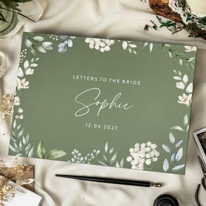 Puede incluir: Una caja rectangular de color verde salvia con un borde floral blanco de hojas y flores. El texto dice "LETTERS TO THE BRIDE", "Sophie" y "12.06.2027" en blanco. La caja está sobre una superficie de tela blanca con un bolígrafo negro al lado.