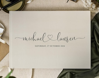 May include: A rectangular guest book with a cream-coloured cover featuring the black printed names michael and lauren connected by a heart symbol, with the text SATURDAY, 27 OCTOBER 2026 printed in a smaller font directly below the names.