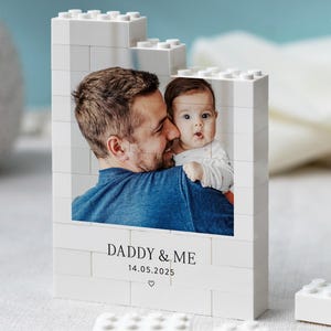 May include: A white rectangular block structure built from interlocking plastic bricks featuring a printed photograph of an adult and a baby. The text DADDY & ME and 14.05.2025 with a small heart icon are printed below the photo.
