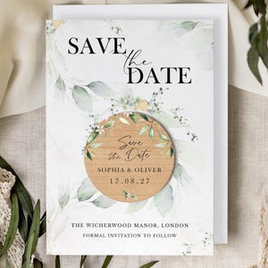 May include: A rectangular Save the Date card featuring green watercolor leaves and black text, with a circular wooden magnet attached to the center. The magnet displays the text Save the Date, Sophia & Oliver, and the date 17.08.27 in black ink.