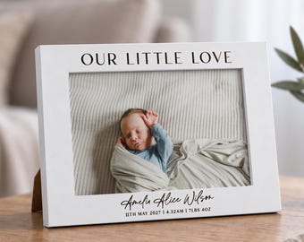 May include: A white rectangular picture frame featuring the black text OUR LITTLE LOVE at the top and the name Amelia Alice Wilson with the birth details 11TH MAY 2027 | 4.32AM | 7LBS 4OZ printed in black at the bottom, displaying a photo of a baby.