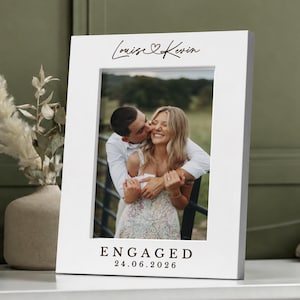 Puede incluir: Un marco de fotos rectangular blanco con los nombres impresos Louise y Kevin junto a un símbolo de corazón en la parte superior. El marco muestra una fotografía de una pareja abrazada e incluye el texto ENGAGED y la fecha 24.06.2026.