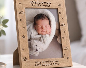 May include: A light brown wooden picture frame featuring the engraved text Welcome to the world at the top and Harry Robert Martins 18th AUGUST 2027 at the bottom. The frame is decorated with small engraved clouds, stars, and rainbows.