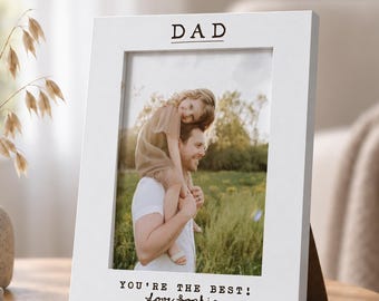 Può includere: Una cornice portafoto rettangolare bianca con la parola DAD stampata in alto e il testo YOU'RE THE BEST! Love Sophia stampato in basso. La cornice contiene una fotografia di una persona che porta un bambino sulle spalle all'aperto.