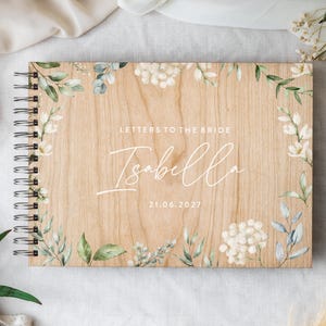 Puede incluir: Un libro de visitas de madera con encuadernación en espiral y acabado de veta de madera clara. La cubierta presenta un borde floral impreso con hojas verdes y flores blancas. El texto dice LETTERS TO THE BRIDE, Isabella y 21.06.2027.