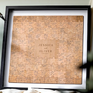 Könnte beinhalten: Ein quadratisches Puzzle-Gästebuch aus Holz in einem schwarzen Rahmen mit weißem Passepartout. Die Puzzleteile sind in der Mitte mit dem Text JESSICA AND OLIVER 08.04.2027 graviert und von einzelnen Teilen mit handschriftlichen Nachrichten in schwarzer Tinte umgeben.