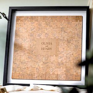 May include: A framed wooden jigsaw puzzle guest book. The light brown puzzle pieces are assembled into a square. The centre of the puzzle is engraved with the names "Olivia and Henry" and a date. Many pieces have handwritten messages.