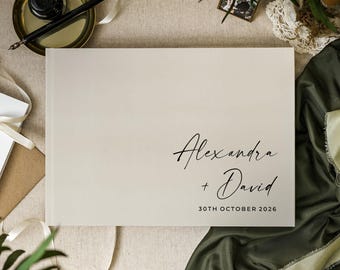 May include: A rectangular off-white guest book featuring the black printed text Alexandra + David and the date 30TH OCTOBER 2026. The cover has a smooth, matte finish and is positioned on a neutral surface with a black ink pen and inkwell.