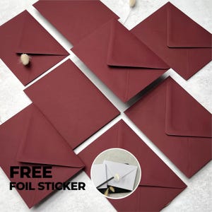 May include: A collection of deep burgundy envelopes arranged in a cluster. One envelope is partially open, revealing a small, fluffy, light-coloured botanical accent. A circular inset shows a white envelope sealed with a gold foil leaf sticker.