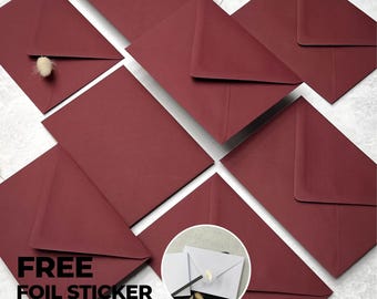 Pode incluir: Uma coleção de envelopes em tom bordô escuro dispostos em grupo. Um envelope está parcialmente aberto, revelando um pequeno detalhe botânico felpudo de cor clara. Um círculo mostra um envelope branco selado com um adesivo de folha dourada.