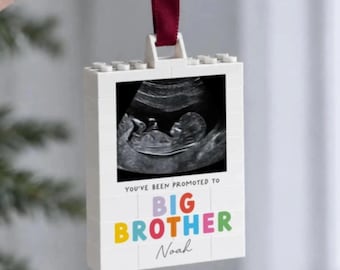 May include: A white rectangular building block ornament featuring a black and white ultrasound image. The text reads YOU'VE BEEN PROMOTED TO BIG BROTHER with the name Noah printed below in script. A dark red ribbon is attached to the top.
