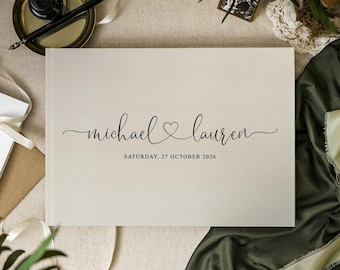 May include: A cream-colored wedding guest book with the names "Michael & Lauren" in elegant script, a heart symbol, and the date "Saturday, 27 October 2026." The book is open, with a pen and inkwell in the background.