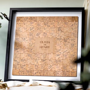 May include: A square wooden jigsaw puzzle guest book framed in black, featuring individual puzzle pieces signed with handwritten messages. The centre piece is engraved with the text OLIVIA and HENRY and the date 08.07.2025 in a serif font.