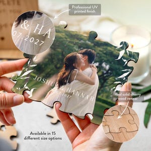 Close-up of the custom UV-printed wooden photo jigsaw puzzle wedding guestbook featuring a photograph of a couple embracing outdoors framed by white botanical leaves and text reading JOSHUA & HANNAH, and 28.07.2027, shown with the matching wooden ins