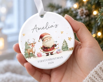 May include: A round white ceramic Christmas ornament featuring a watercolor illustration of Santa Claus and a reindeer with gifts and trees. The text reads Amelia's FIRST CHRISTMAS 2026. The ornament hangs from a white ribbon for display.